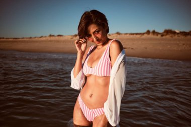Seksi, güzel Latin kız, ıssız bir sahilde banyo yapıyor pembe bir bikini ve pembe güneş gözlüğü takıyor.