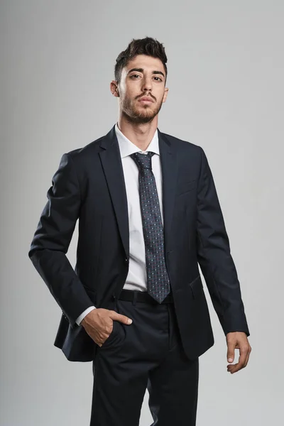 Sakallı, koyu mavi takım elbiseli genç adam sağ elini pantolonunun cebine sokup kameraya bakıyor..