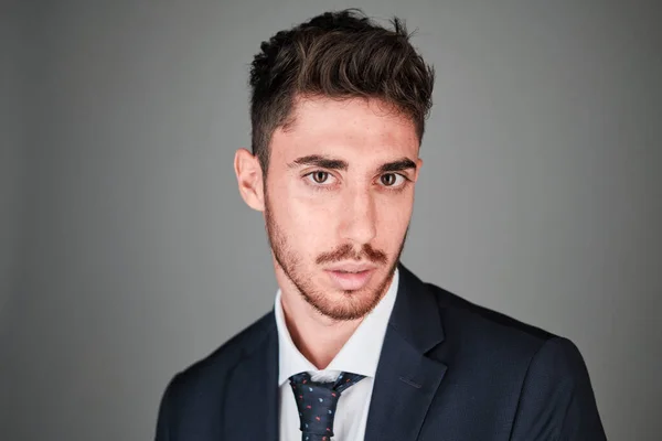 Genç adam, koyu mavi bir takım elbise ve kırmızı ve açık mavi noktalı koyu mavi bir kravat takıyor..