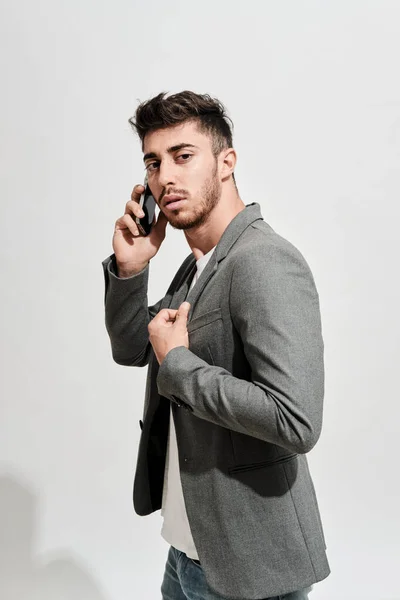 Kot pantolon, beyaz tişört ve gri ceketli genç bir adam cep telefonuyla konuşurken beyaz bir duvarın önünde yürüyordu.