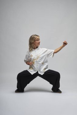 Olgun sarışın kadın beyaz arka planda Chi kung ve Tai Chi çalışıyor. Geleneksel beyaz Çin Tai Chi ceketi, siyah pantolon ve ying yang sembollü siyah ayakkabılar giyiyor.