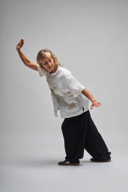 Olgun sarışın kadın beyaz arka planda Chi kung ve Tai Chi çalışıyor. Geleneksel beyaz Çin Tai Chi ceketi, siyah pantolon ve ying yang sembollü siyah ayakkabılar giyiyor.