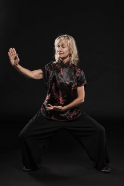 Siyah arka planda Tai Chin Chen stili pratik yapan olgun sarışın bir kadın. Kırmızı madalyalı, siyah pantolonlu ve ying yang sembollü geleneksel ayakkabılı trajik bir Çinli siyah ceket giyiyor.