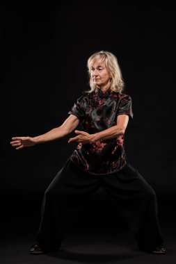 Siyah arka planda Tai Chin Chen stili pratik yapan olgun sarışın bir kadın. Kırmızı madalyalı, siyah pantolonlu ve ying yang sembollü geleneksel ayakkabılı trajik bir Çinli siyah ceket giyiyor.