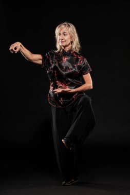 Siyah arka planda Tai Chin Chen stili pratik yapan olgun sarışın bir kadın. Kırmızı madalyalı, siyah pantolonlu ve ying yang sembollü geleneksel ayakkabılı trajik bir Çinli siyah ceket giyiyor.