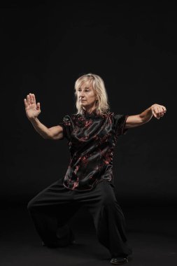 Siyah arka planda Tai Chin Chen stili pratik yapan olgun sarışın bir kadın. Kırmızı madalyalı, siyah pantolonlu ve ying yang sembollü geleneksel ayakkabılı trajik bir Çinli siyah ceket giyiyor.