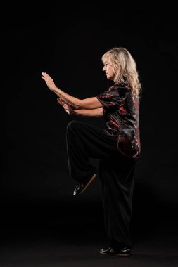 Siyah arka planda Tai Chin Chen stili pratik yapan olgun sarışın bir kadın. Kırmızı madalyalı, siyah pantolonlu ve ying yang sembollü geleneksel ayakkabılı trajik bir Çinli siyah ceket giyiyor.