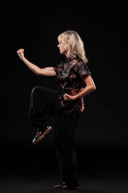 Siyah arka planda Tai Chin Chen stili pratik yapan olgun sarışın bir kadın. Kırmızı madalyalı, siyah pantolonlu ve ying yang sembollü geleneksel ayakkabılı trajik bir Çinli siyah ceket giyiyor.