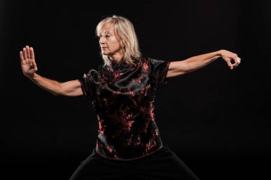 Siyah arka planda Tai Chin Chen stili pratik yapan olgun sarışın bir kadın. Kırmızı madalyalı, siyah pantolonlu ve ying yang sembollü geleneksel ayakkabılı trajik bir Çinli siyah ceket giyiyor.