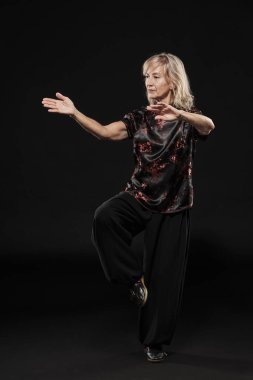 Siyah arka planda Tai Chin Chen stili pratik yapan olgun sarışın bir kadın. Kırmızı madalyalı, siyah pantolonlu ve ying yang sembollü geleneksel ayakkabılı trajik bir Çinli siyah ceket giyiyor.