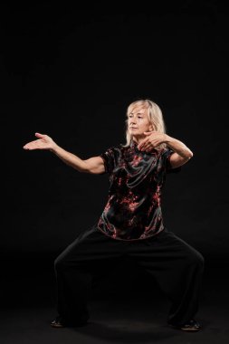 Siyah arka planda Tai Chin Chen stili pratik yapan olgun sarışın bir kadın. Kırmızı madalyalı, siyah pantolonlu ve ying yang sembollü geleneksel ayakkabılı trajik bir Çinli siyah ceket giyiyor.