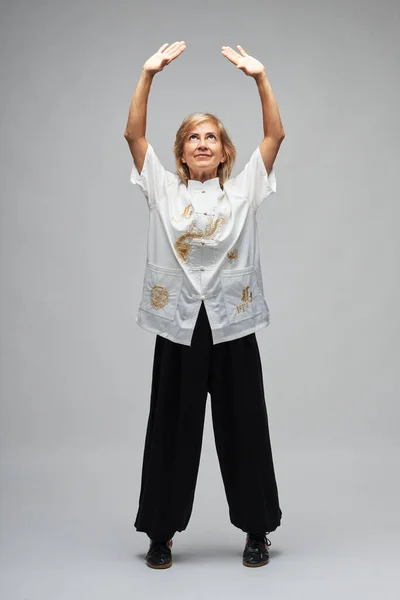 Olgun sarışın kadın beyaz arka planda Chi kung ve Tai Chi çalışıyor. Geleneksel beyaz Çin Tai Chi ceketi, siyah pantolon ve ying yang sembollü siyah ayakkabılar giyiyor.