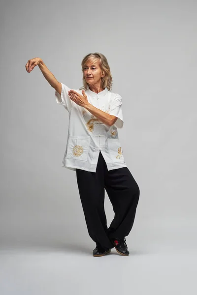 Olgun sarışın kadın beyaz arka planda Chi kung ve Tai Chi çalışıyor. Geleneksel beyaz Çin Tai Chi ceketi, siyah pantolon ve ying yang sembollü siyah ayakkabılar giyiyor.