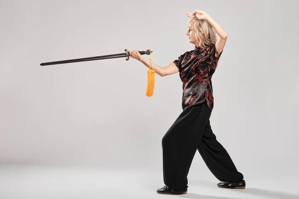 Beyaz arka planda Tai Chin 'e kılıç çeken olgun sarışın bir kadın. Geleneksel Çin siyah ceketi, kırmızı Çin süslemesi, siyah pantolon ve Ying Yang sembollü Çin ayakkabısı giyiyor.