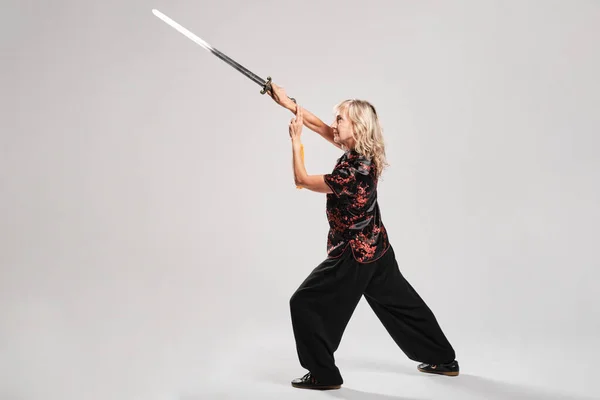 Mature blonde woman practicing Tai Chin with sword in a white background wearing a traditional chinese black jacket with red chinese decoration, black trousers and chinese shoes with ying yang symbol