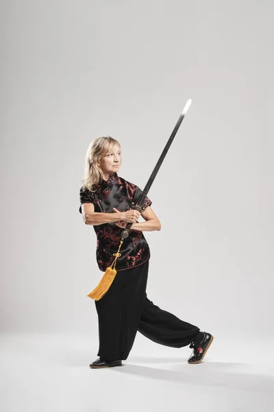 Beyaz arka planda Tai Chin 'e kılıç çeken olgun sarışın bir kadın. Geleneksel Çin siyah ceketi, kırmızı Çin süslemesi, siyah pantolon ve Ying Yang sembollü Çin ayakkabısı giyiyor.