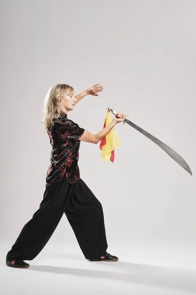 Beyaz arka planda Tai Chin 'e kılıç çeken olgun sarışın bir kadın. Geleneksel Çin siyah ceketi, kırmızı Çin süslemesi, siyah pantolon ve Ying Yang sembollü Çin ayakkabısı giyiyor.