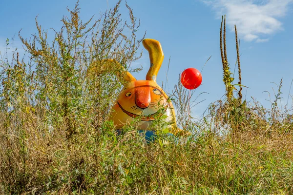 Huge funny plastic rabbit with ears and a balloon in the bushes against the blue sky