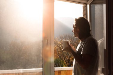 Elinde bir kupa sıcak çayla yetişkin bir adam ayakta durup dağlara bakar..