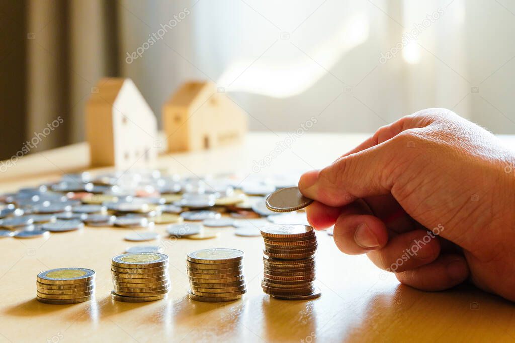 Men hand put coin at increase stack with blurred house at the background and natural light. Investment or saving for dream home ownership concept. Property loan and mortgage.
