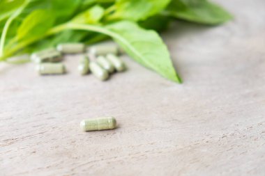 Kahverengi ahşap yüzeydeki kapsüldeki bitkisel ilaçlara seçici bir şekilde odaklan, arka planda yeşil fesleğen yaprağı bitkisi olsun. Sağlık ve tıbbi yaşam tarzı kavramı.