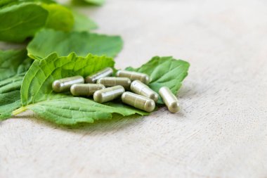 Kahverengi ahşap yüzeydeki kapsüldeki bitkisel ilaçlara seçici bir şekilde odaklan, arka planda yeşil fesleğen yaprağı bitkisi olsun. Sağlık ve tıbbi yaşam tarzı kavramı.