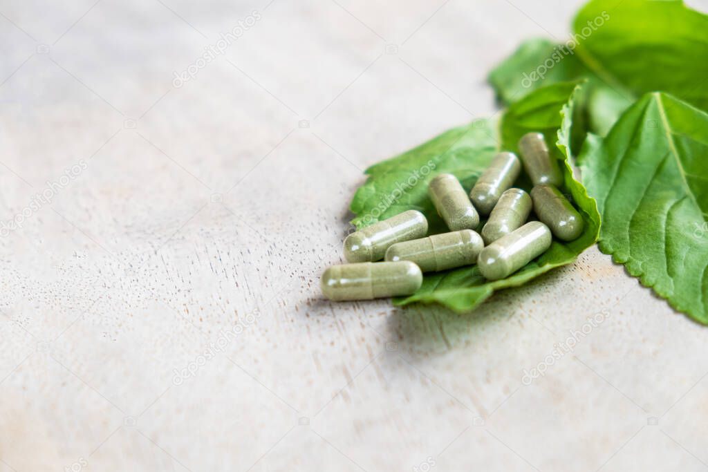 Selective focus at Herbal medicine in capsule on brown wood surface, with green  holy basil leaf herb at the background. Healthcare and medical lifestyle concept.