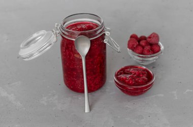 Cam bir kavanozda ahududu reçeli ve fincanda olgun ahududular ve gri bir arka planda bir kaşık.