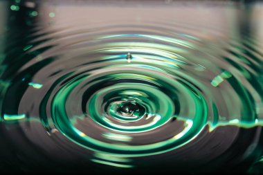 Bir su damlacığının detaylı yakın çekimi, zümrüt yeşili bir yüzeyde, üzerinde bir damla bulunan, hassas, eşmerkezli bir dalgalanma deseni yaratıyor..