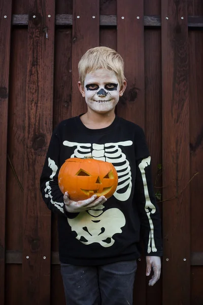 Happy young blond hair boy with skeleton costume holding jack o lantern. Halloween. Trick or treat. Outdoors portrait over wooden background