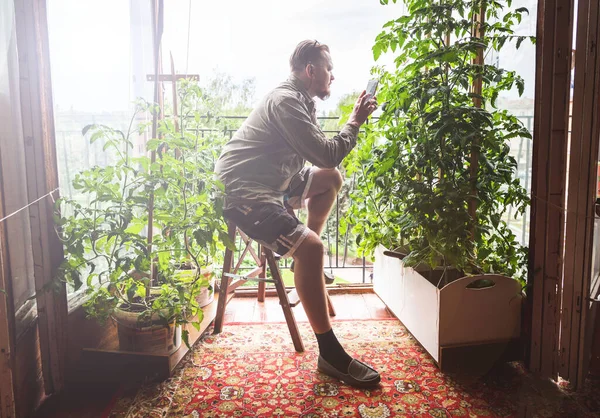 The gardener works in a mini-garden on the balcony. A man photographs with a smartphone his plants in a city apartment.