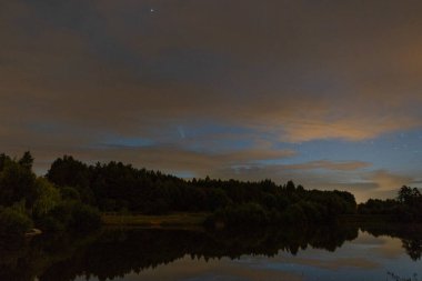 Akşam manzarası: Bulutlarda yıldızlı bir gökyüzü ve güneş battıktan hemen sonra bir orman gölü üzerinde Neowise kuyruklu yıldızı. Uzun gece manzarası. 