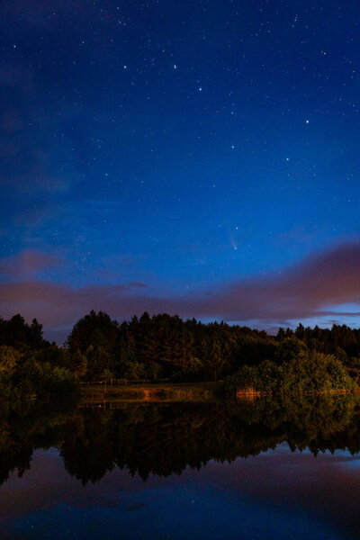 Evening landscape: a starry sky in the clouds and a comet Neowise over a forest lake just after sunset.Long exposure night landscape. The constellation Ursa Major and a comet in the sky above trees and lake.