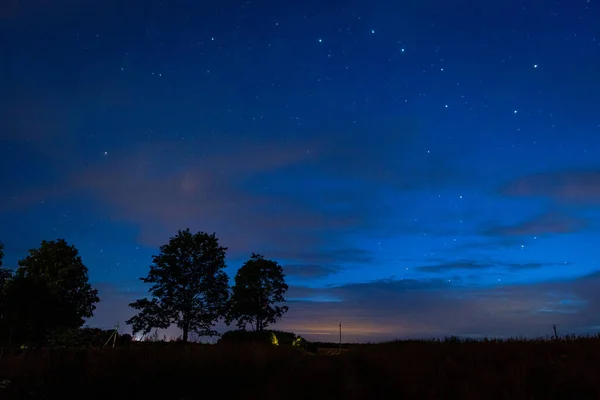 The constellation Ursa Major and a comet in the sky above trees and a village house. Evening landscape: a starry sky in the clouds and a comet Neowise over a forest lake just after sunset. Long exposure night landscape.