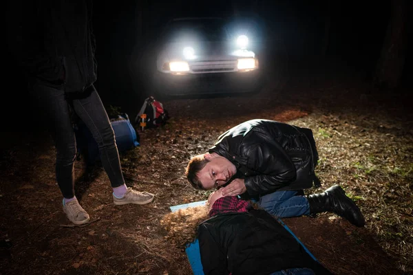 Field First Aid Training. A man is checked to see if a woman lying on the ground is breathing, another woman is standing nearby. Night scene in the headlights of a car.