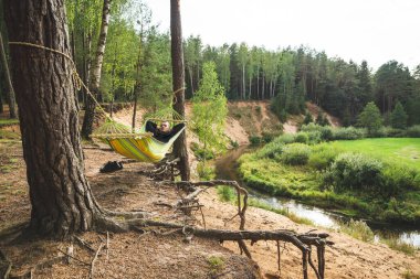 Dik bir nehir kıyısındaki bir hamakta dinlenen bir adam. Genel plan