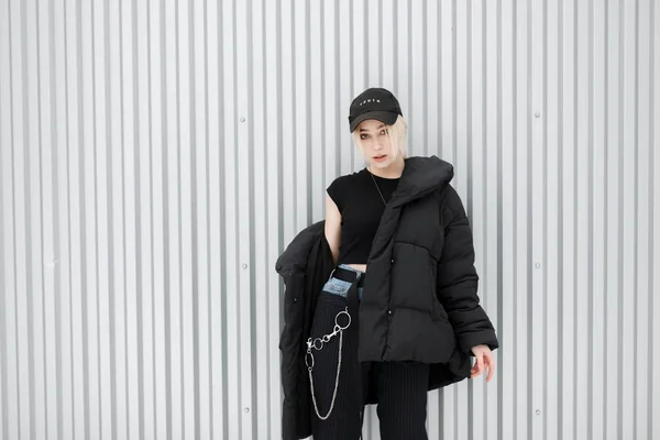 金属製の壁の近くのキャップとファッショナブルな現代冬黒ジャケットでスタイリッシュな美しいファッション モデルの女の子 — ストック写真