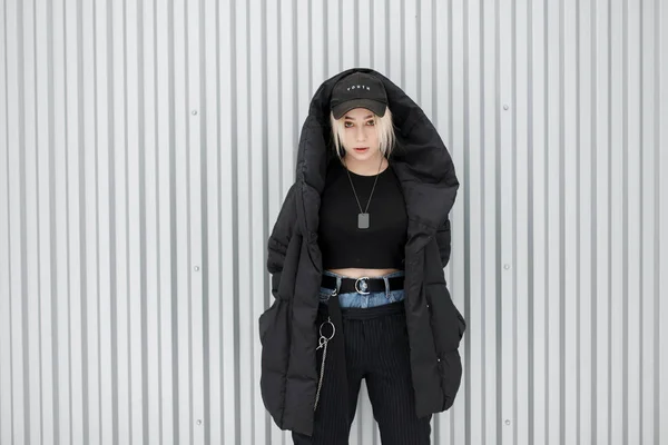 时尚漂亮的年轻女孩在黑色时尚的冬季夹克与一个时髦的帽子摆在金属墙附近 — 图库照片