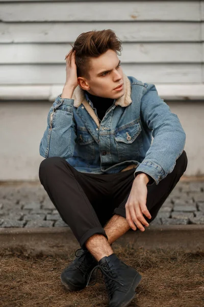 beautiful fashion model a man with a haircut in a fashionable denim jacket with black pants and shoes sitting near a wooden wall