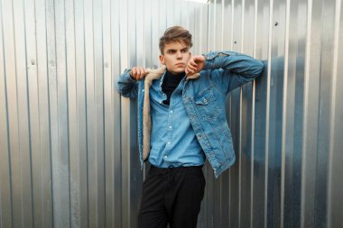 Şık yakışıklı genç bir metal duvar yakınında sokakta poz kot gömlek moda kot ceketli bir saç modeli ile