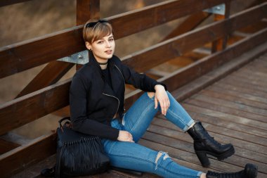 Siyah şık çanta ve bot ile siyah ceketli güzel genç kadın bir ahşap köprü üzerinde oturur