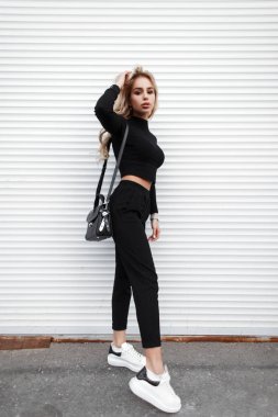 Şık genç model kadın beyaz spor ayakkabı metal bir duvara yakın poz ile şık siyah giysili bir deri çanta