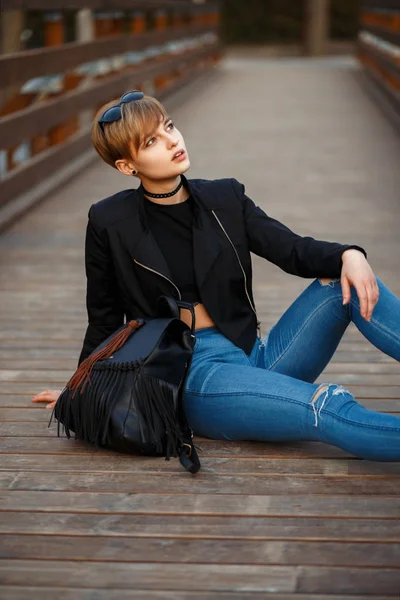 Fashionable young model in black stylish clothes with a jacket, jeans and boots with a leather bag sitting on a wooden bridge on the street