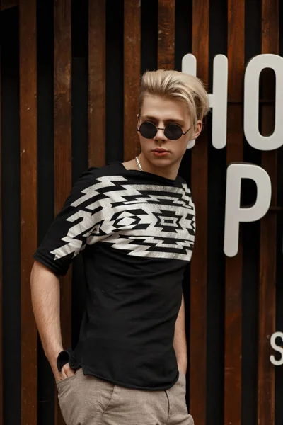 Handsome fashion model man with a hairstyle in vintage round sunglasses in a black stylish T-shirt posing near a wooden wall in white letters