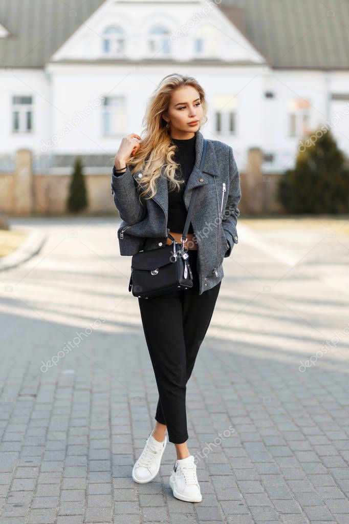 Beautiful young stylish girl with a bag in a fashion gray coat, black pants and white sneakers in the street on a sunny day