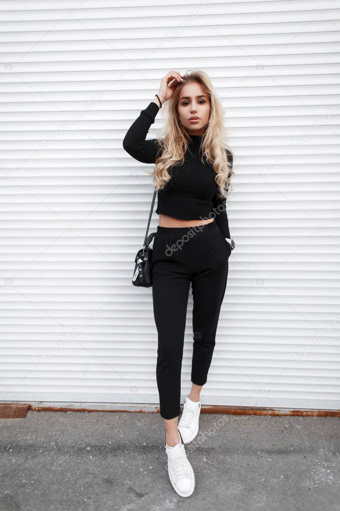 Young beautiful fashion model girl with a handbag in a black T-shirt, pants and white sneakers poses near a white metal wall