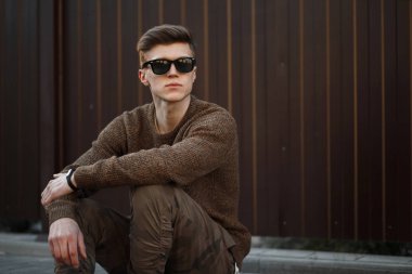 Yakışıklı genç hippi erkek güneş gözlüğü metal duvar yakınında bir sokakta oturan moda giysileri