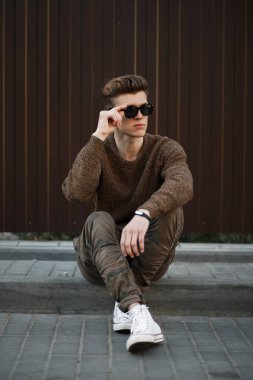 Askeri pantolon ve beyaz spor ayakkabı ile şık giysiler içinde gözlüklü genç moda model adam metal duvar yakınında sokakta oturuyor. 