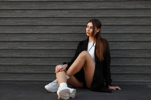 木製の壁のそばに座ってファッショナブルな靴と黒いコートで美しいセクシーなモデルの女性 — ストック写真