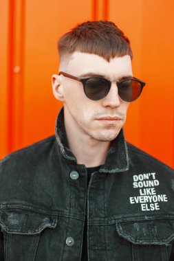 Şık yakışıklı genç moda siyah denim giysileri turuncu bir arka plan üzerinde güneş gözlüklü bir saç modeli ile